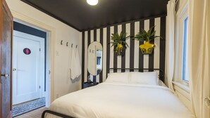 A bedroom with a black and white striped wall, a bed with white bedding, two potted plants, a round mirror, and a door with a female restroom symbol.