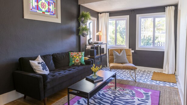 A living room with a dark-colored sofa, a coffee table, a patterned rug, a bookshelf, and a window with curtains.