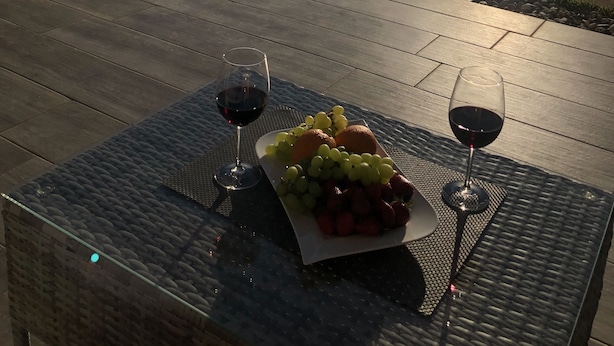Un área de estar al aire libre con piscina, una mesa con una bandeja de frutas y copas de vino, y vistas al océano y a las montañas.