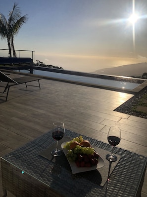 Un área de estar al aire libre con piscina, una mesa con una bandeja de frutas y copas de vino, y vistas al océano y a las montañas.