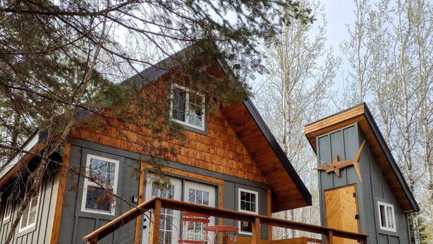 A cabin with a wooden deck, a staircase leading to a porch, and a stack of firewood outside. - House of the Grouse -  Charming  River-side Cabin and Sauna!  (Grand Marais)