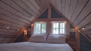 A bedroom with a wooden sloped ceiling, a large bed with white bedding, and two windows with curtains. - House of the Grouse -  Charming  River-side Cabin and Sauna!  (Grand Marais)