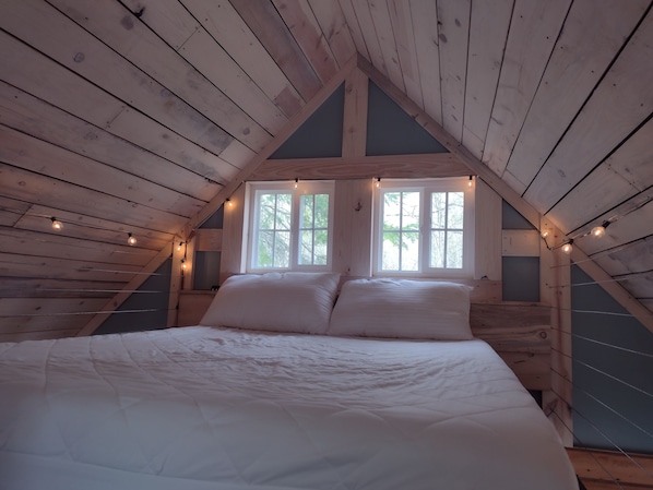 Une chambre avec un plafond en pente en bois, un grand lit avec une literie blanche et deux fenêtres avec des rideaux.