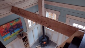 A high-ceilinged room with exposed wooden beams, a fireplace, and a colorful painting on the wall. - House of the Grouse -  Charming  River-side Cabin and Sauna!  (Grand Marais)