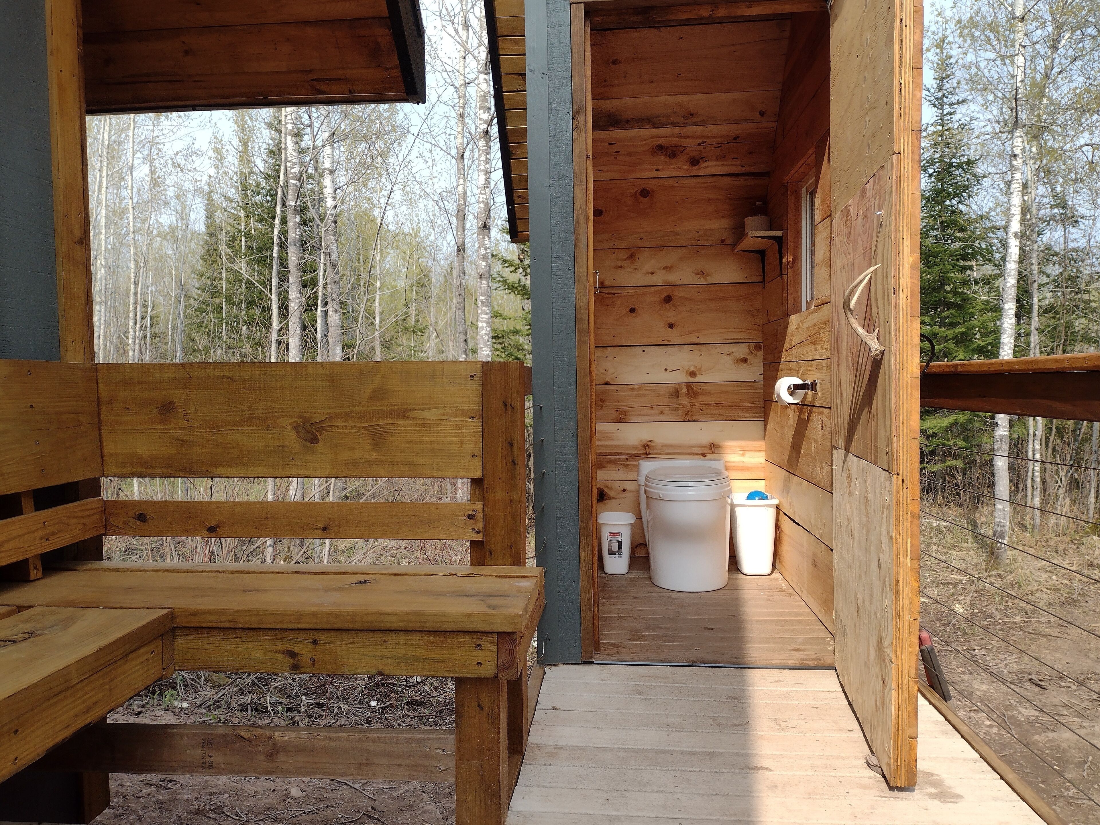 Una cabaña de madera con un banco, un inodoro y un cubo de basura.