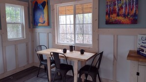 A dining area with a wooden table and chairs, large windows, and two paintings on the walls. - House of the Grouse -  Charming  River-side Cabin and Sauna!  (Grand Marais)