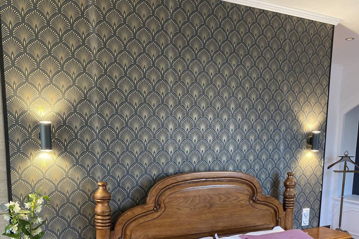 Une chambre à coucher bien aménagée, avec un papier peint à motifs, une tête de lit en bois, une table de chevet agrémentée d’une plante et une commode.