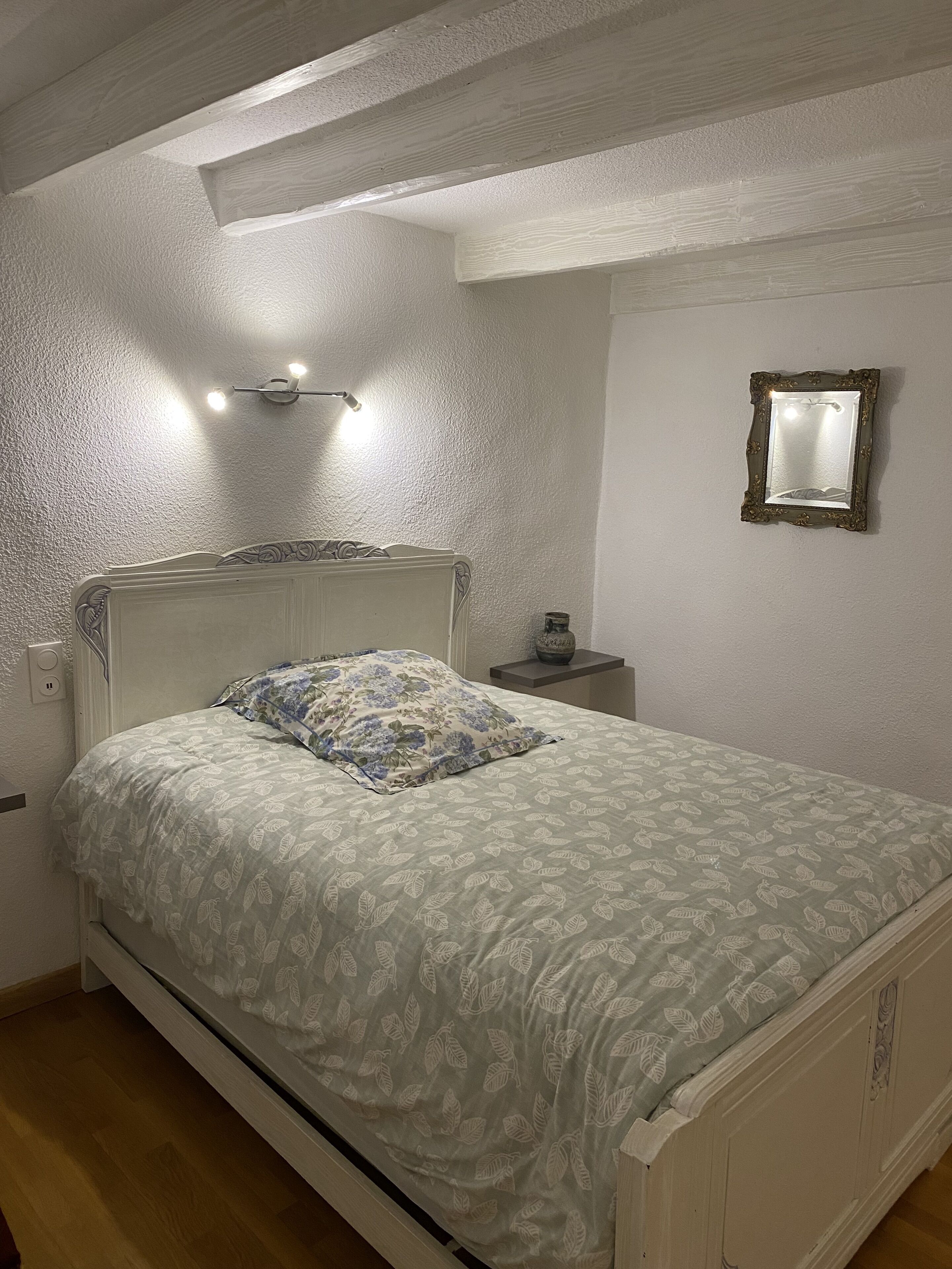 Un dormitorio con una cama, una mesita de noche, un espejo en la pared y una lámpara en el techo.