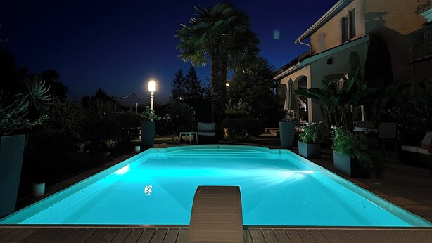 Een zwembad in de nacht met een houten terras op de voorgrond en een gebouw met balkon op de achtergrond.