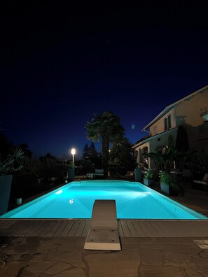 Une piscine la nuit, avec une terrasse en bois au premier plan et un bâtiment doté d’un balcon en arrière-plan.