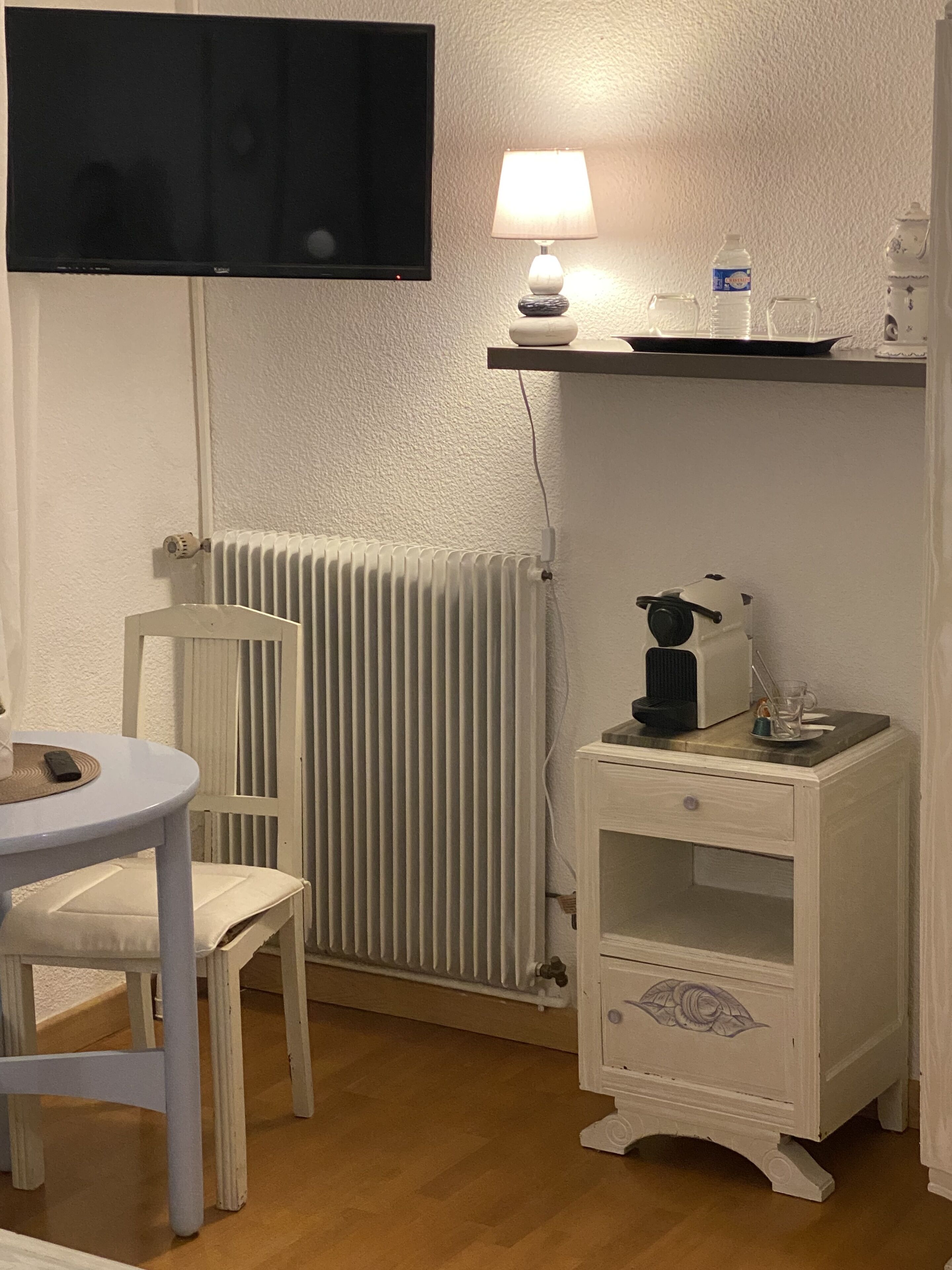 A room with a white radiator, a small white table with a chair, a white cabinet with a drawer, a lamp, a television mounted on the wall, and a shelf with a bottle and glasses.