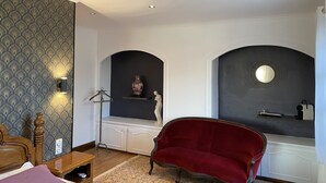 Een kamer met behang met patroon, een rode fluweelstoel, een houten bed, een tafeltje, een decoratieve vaas en een spiegel aan de muur.
