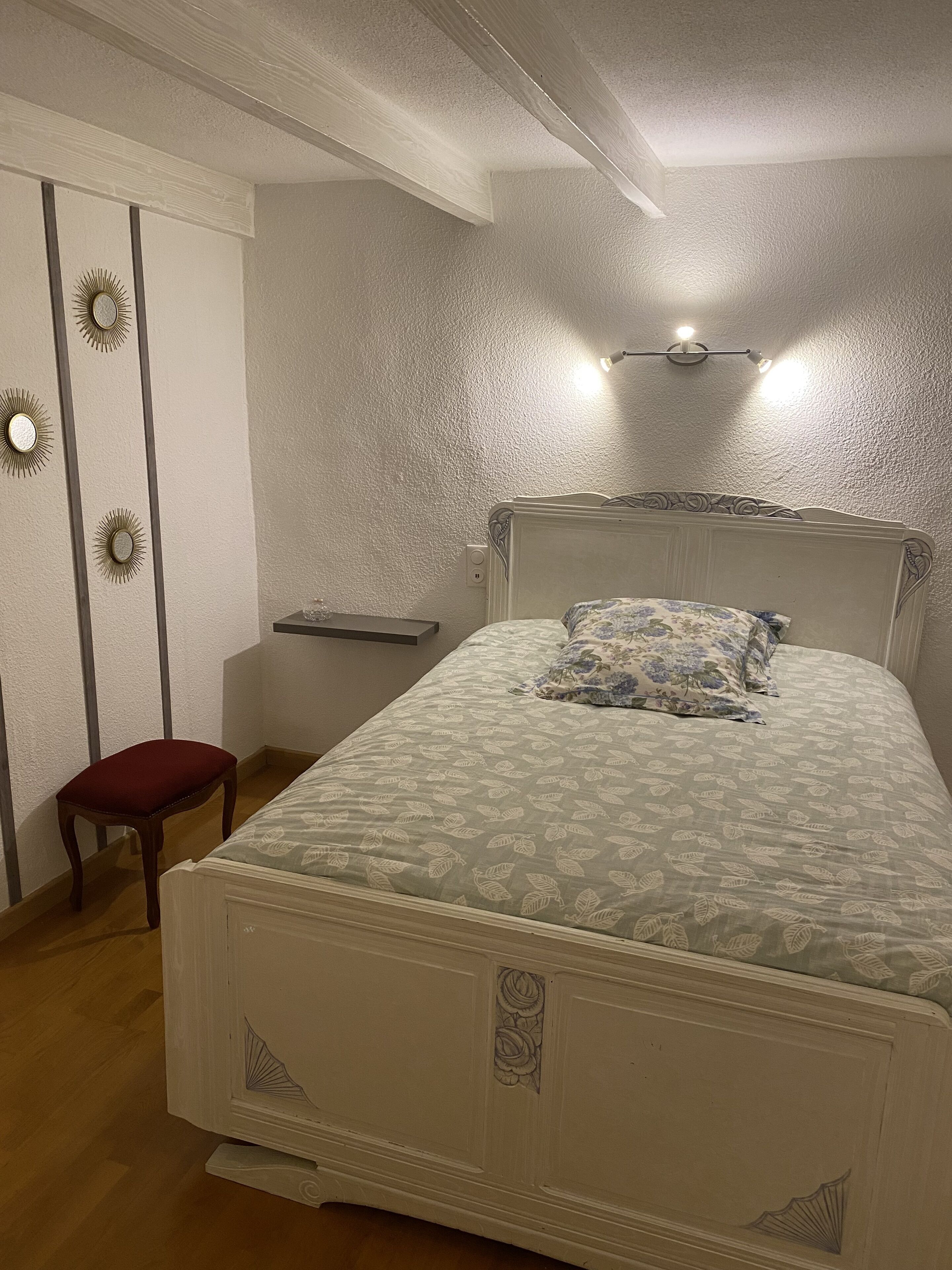 Een slaapkamer met een groot bed, een klein rode krukje, een nachtkastje en wandlampen.
