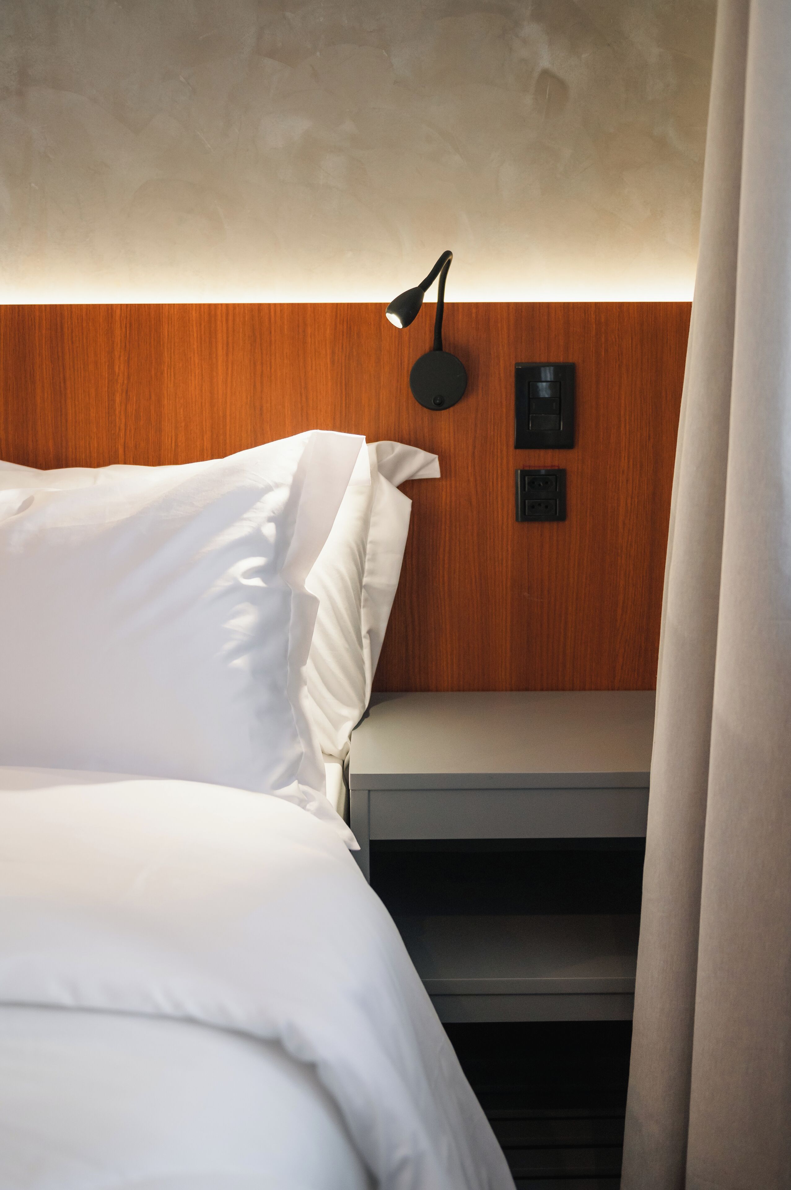 Ein ordentlich bezogenes Bett mit weißen Leinen, einem hölzernen Kopfteil, einer Wandlampe und einer Steckdose.