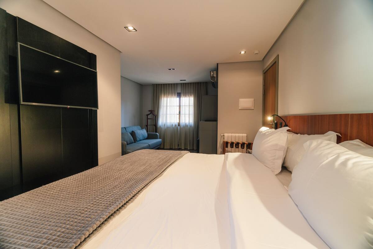 Habitación de hotel con una cama grande, un televisor de pantalla plana en la pared, un sofá azul y una ventana con cortinas.