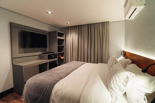 A modern hotel room with a large bed, a flat-screen TV mounted on the wall, a desk with a computer, and a built-in air conditioning unit.