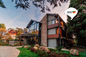 A modern multi-story building with large glass windows, a stone facade, and a landscaped garden with rocks and shrubs. - 249 DESIGN HOTEL (Gramado)