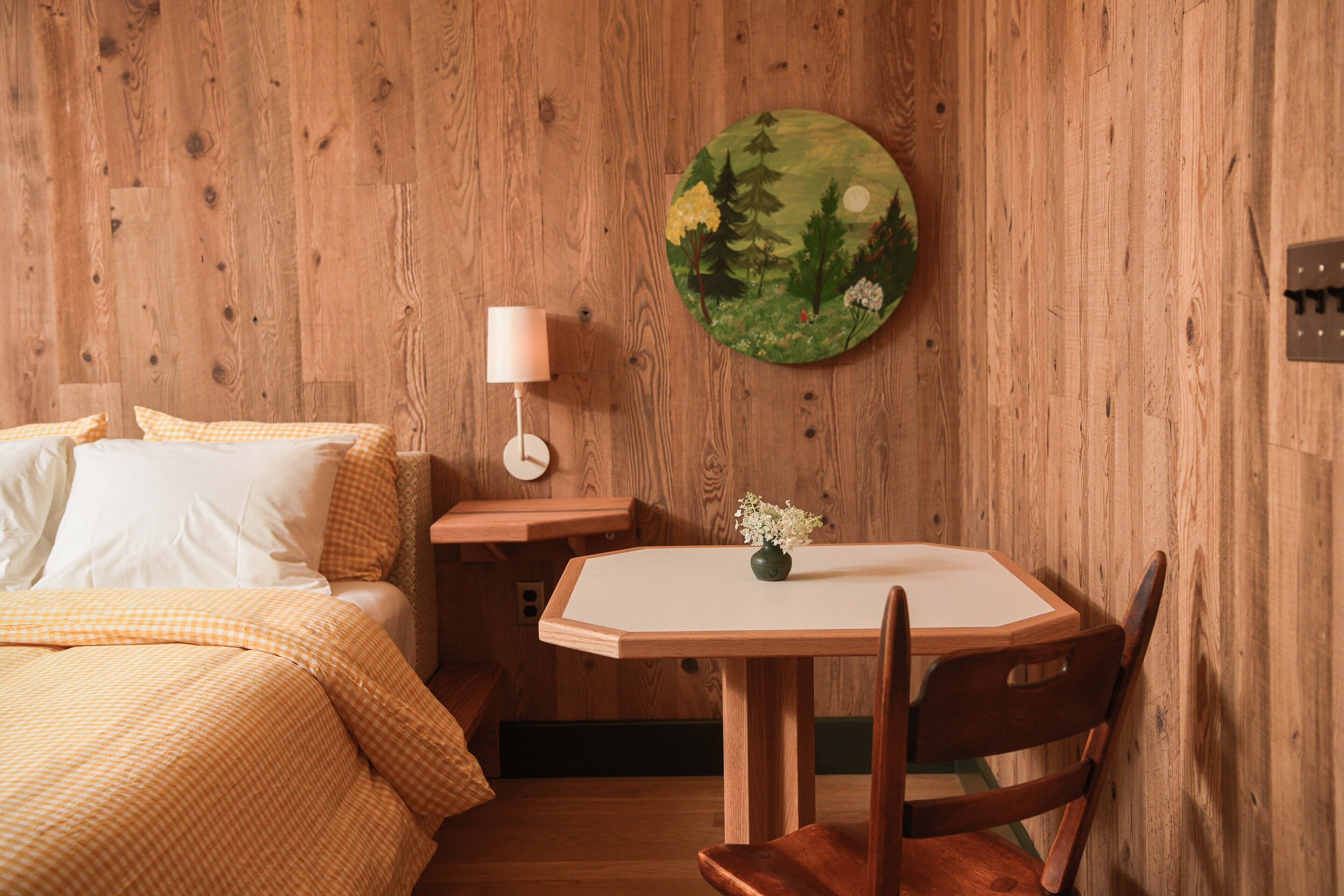 A room with a wooden wall, a bed with yellow bedding, a bedside table with a lamp, and a round painting of a forest scene.