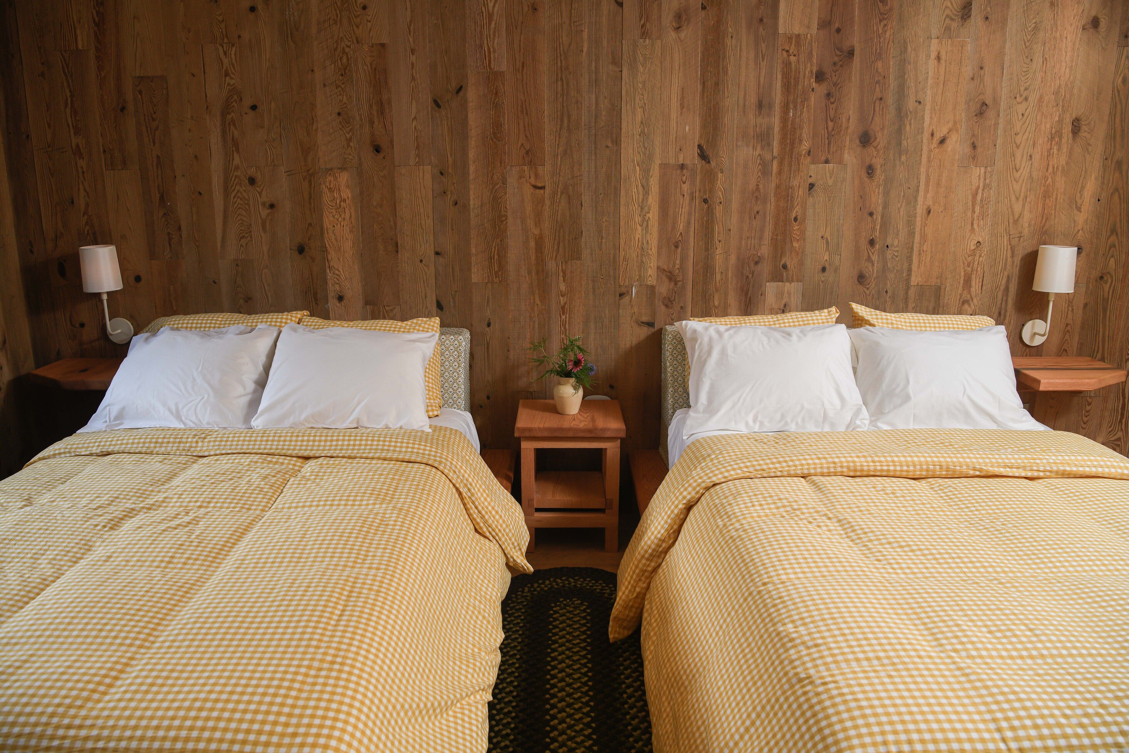 A room with two beds, each with a checkered bedspread and white pillows, against a wooden wall. There are two bedside tables with lamps, and a small table with a vase and plant in the center.