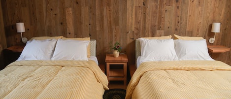 Una habitación con dos camas, cada una con cubrecama a cuadros y almohadas blancas, frente a una pared de madera. Hay dos mesitas de noche con lámparas, y una pequeña mesa con un florero y una planta en el centro.