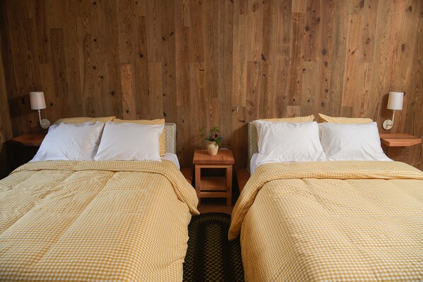 Una habitación con dos camas, cada una con cubrecama a cuadros y almohadas blancas, frente a una pared de madera. Hay dos mesitas de noche con lámparas, y una pequeña mesa con un florero y una planta en el centro.