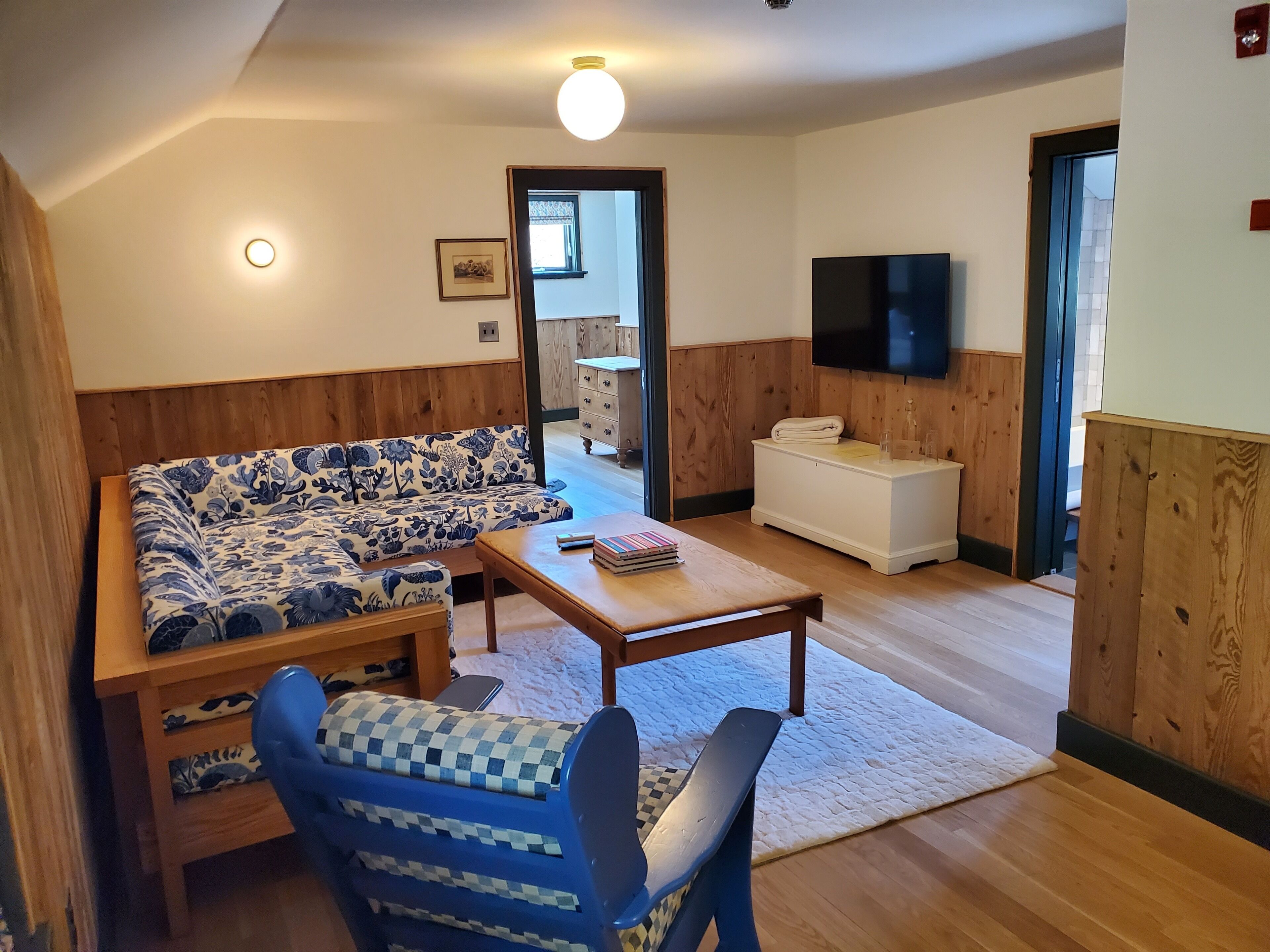 A cozy living room with a patterned sofa, a wooden coffee table, and a blue armchair. A flat-screen TV is mounted on the wall, and a wooden cabinet is visible in the background.