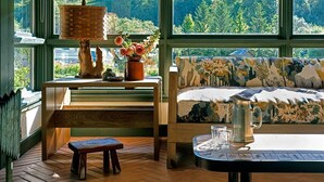Ein gemütlicher Raum mit einer hölzernen Bank, einem Tisch mit einem Krug und einer Couch mit gemusterten Kissen. Große Fenster mit gelben Jalousien bieten Blick ins Grüne.
