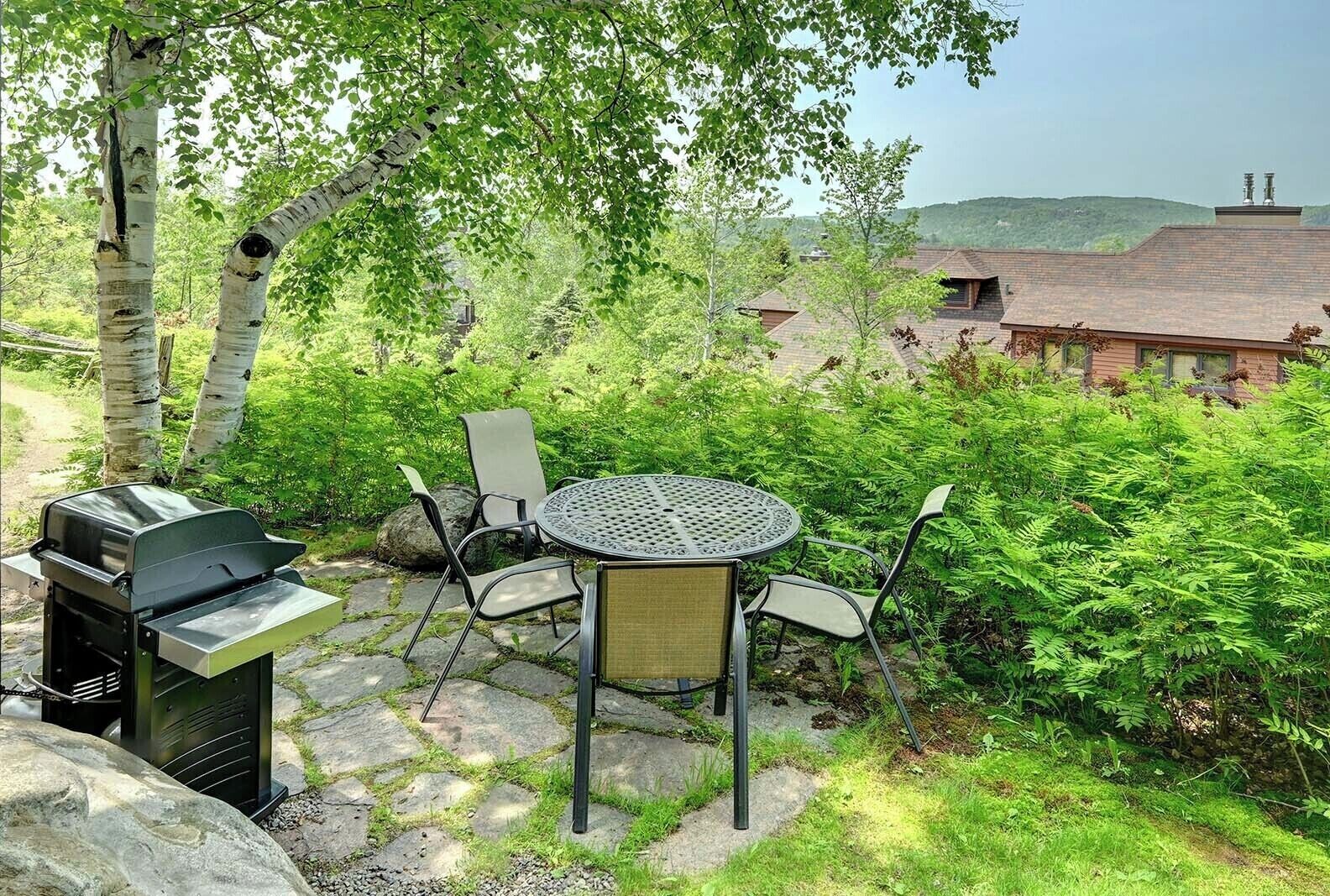 Una terrazza esterna con un tavolo rotondo e sedie, immersa nel verde e con vista su una casa sullo sfondo.
