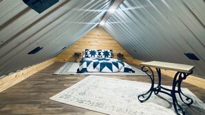 Ein Dachzimmer mit hohen Decken und geneigter Schräge, Holzboden, einem Bett mit gemustertem Überwurf, einem kleinen Tisch und einer Wandleuchte.