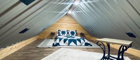 Ein Dachzimmer mit hohen Decken und geneigter Schräge, Holzboden, einem Bett mit gemustertem Überwurf, einem kleinen Tisch und einer Wandleuchte.