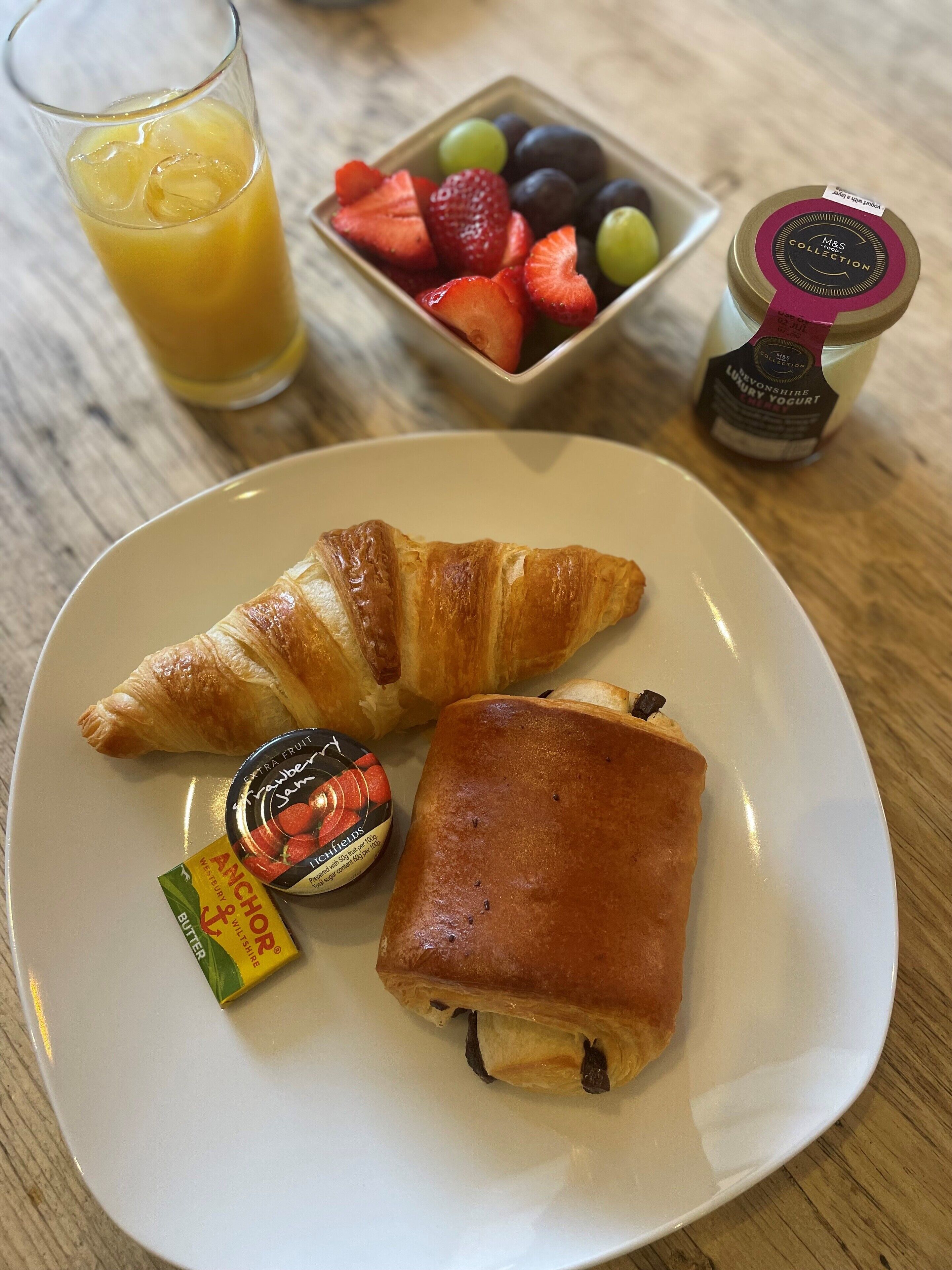 A plate with a croissant and a square pastry, a glass of orange juice, a bowl of mixed berries, and a jar of yogurt.