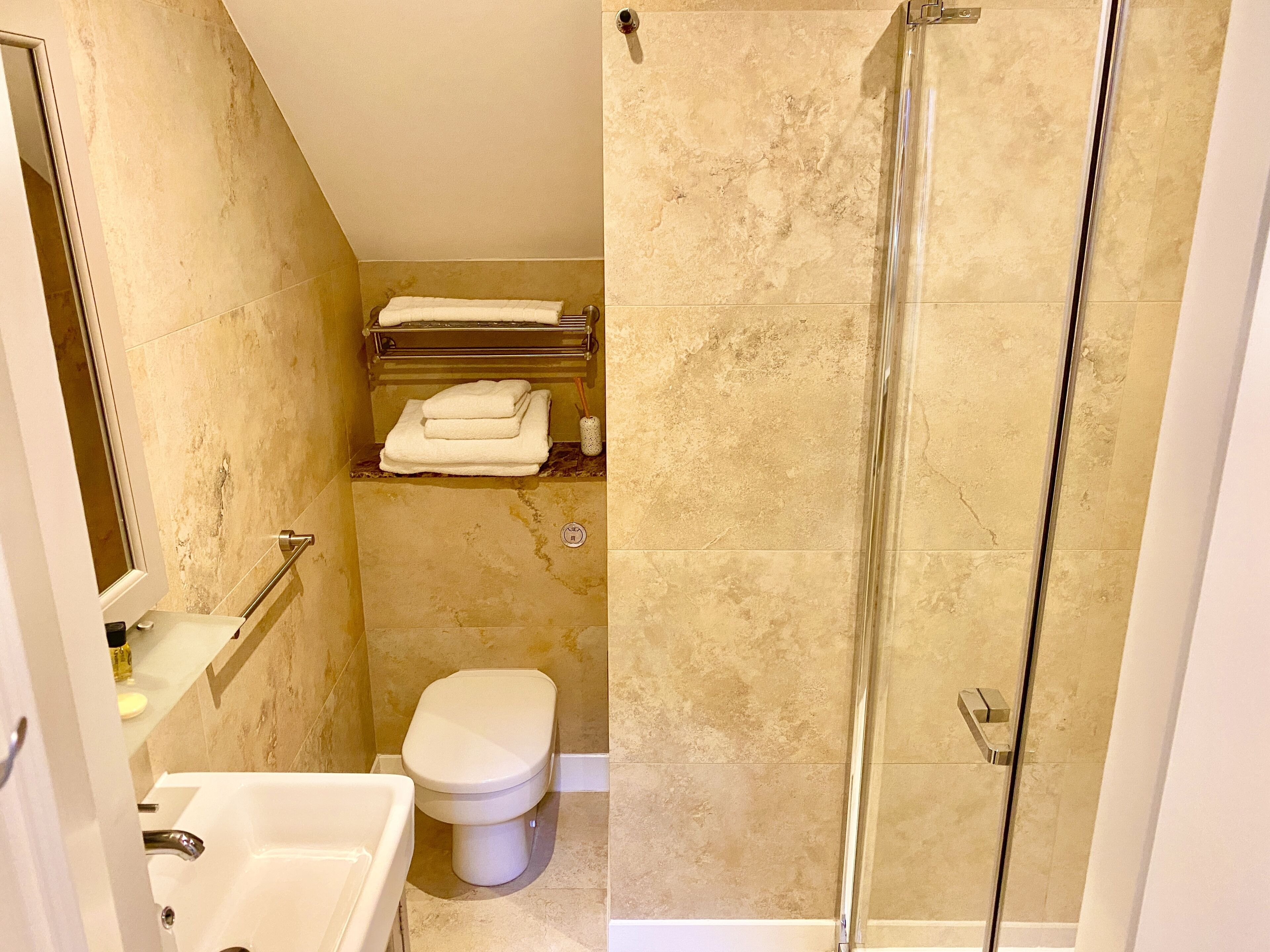 A bathroom with a glass shower enclosure, a toilet, a sink, and a shelf with towels.