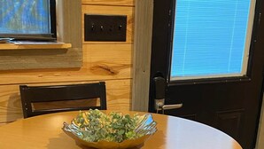 Ein runder Holztisch mit einer Schale grüner Pflanzen, zwei Holzstühle, eine schwarze Tür und ein Fenster mit Jalousien.