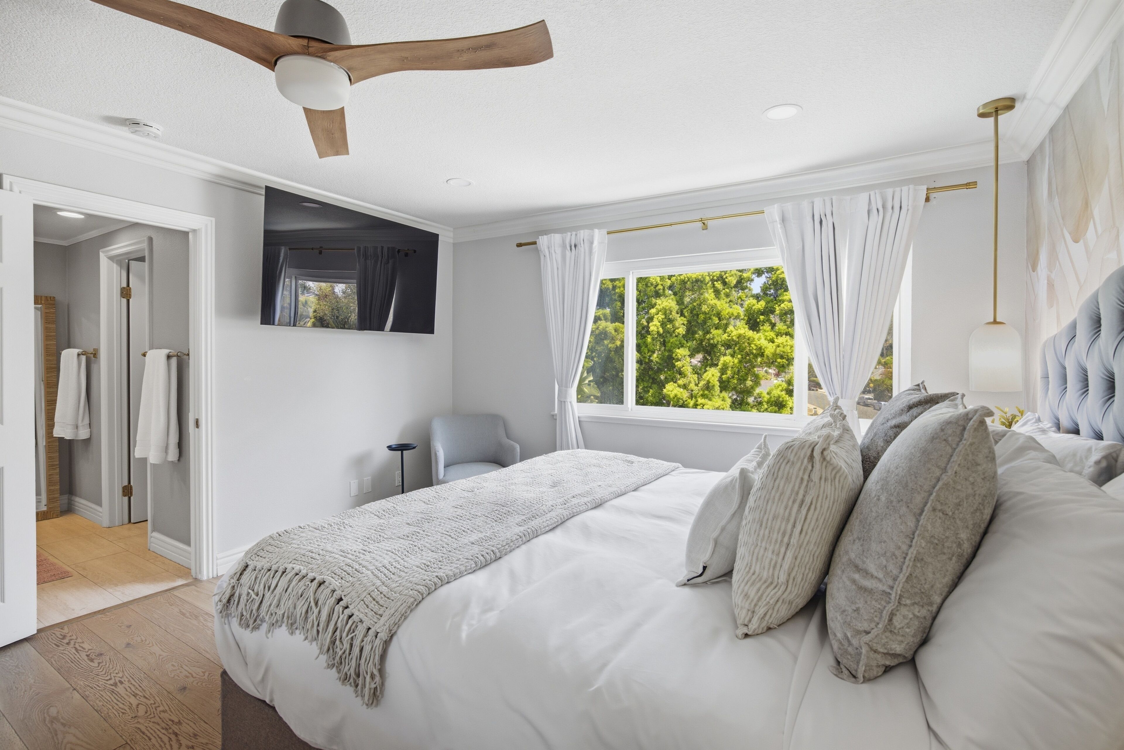 Une chambre à coucher avec un lit, un ventilateur de plafond, une fenêtre avec des rideaux et une vue sur les arbres.