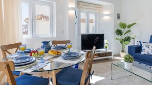 A modern living room with a glass dining table, blue upholstered chairs, a flat-screen TV, and a blue sofa.
