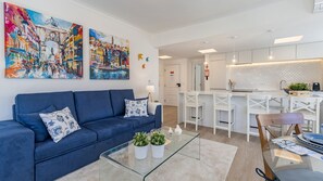 A modern living room with a blue sofa, two paintings, a glass coffee table with plants, and a view into the kitchen area.