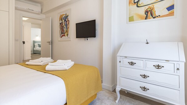 Ett hotellrum med en säng, en vit byrå med utsmyckade handtag, en platt-TV som är monterad på väggen och konstverk ovanför byrån.