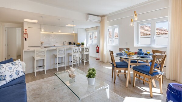A modern kitchen with white cabinetry, a dining area with a glass table, and a living room with a blue sofa.