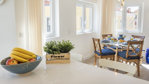 Comedor con una mesa dispuesta para dos, un frutero y una planta en maceta con la etiqueta 'Flower'.