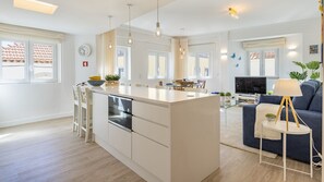 A modern kitchen with a central island, dining area with chairs, a sofa, and a TV.