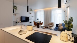 Una cocina moderna con cocina por inducción negra, cafetera y tostadora. Hay un comedor con mesa y sillas, y un sofá en la sala.