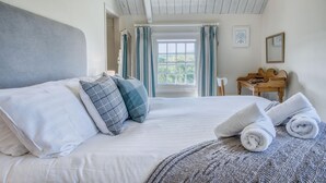 A neatly made bed with white linens and gray pillows, a wooden nightstand with a lamp, and a view of greenery through a window. - Dollars Cottage - 4 Bedroom Cottage - Llangennith (Swansea)