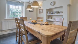 A wooden dining table with chairs, a window with blinds, a clock, and shelves with decorative items. - Dollars Cottage - 4 Bedroom Cottage - Llangennith (Swansea)