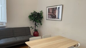 A room with a grey sofa, a wooden table with four chairs, a red potted plant, and a framed picture on the wall.