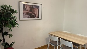 A dining area with a wooden table, two white chairs, a framed picture on the wall, and a red vase with green leaves.
