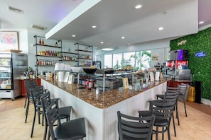 A modern bar area with a granite countertop, bar stools, shelves with bottles, a refrigerator, and a green wall with plants.