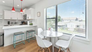 A modern kitchen with white cabinets, a stainless steel microwave, and a red pendant light. There is a round table with white chairs and a window with blinds.