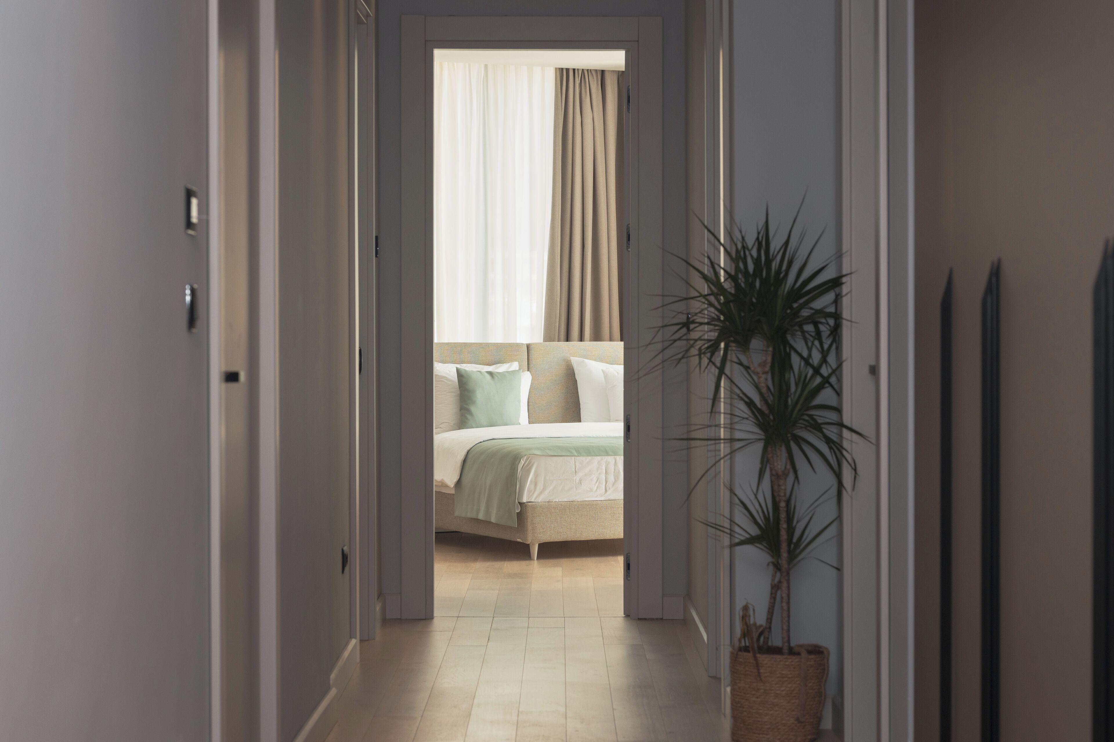 A hallway leading to a bedroom with a bed, a potted plant, and a view of the outside through a window with curtains.