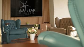 Un soggiorno moderno con un divano blu, un divano beige e un tavolino in legno. In parete, un televisore mostra il logo del 'Sea Star Condo Hotel'.