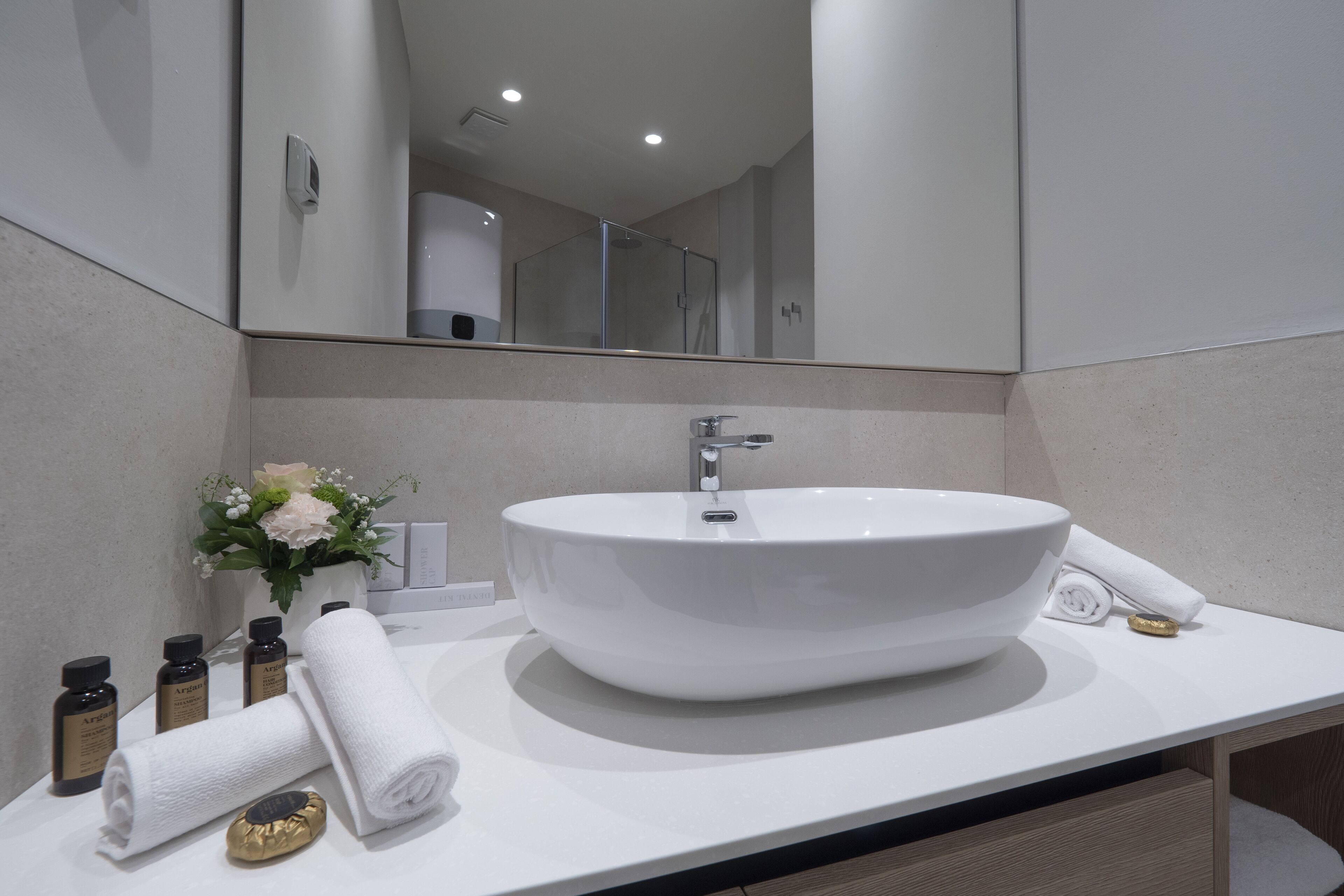 A modern bathroom with a large white sink, a mirror, and a small vase with flowers. There are towels and toiletries on the counter.